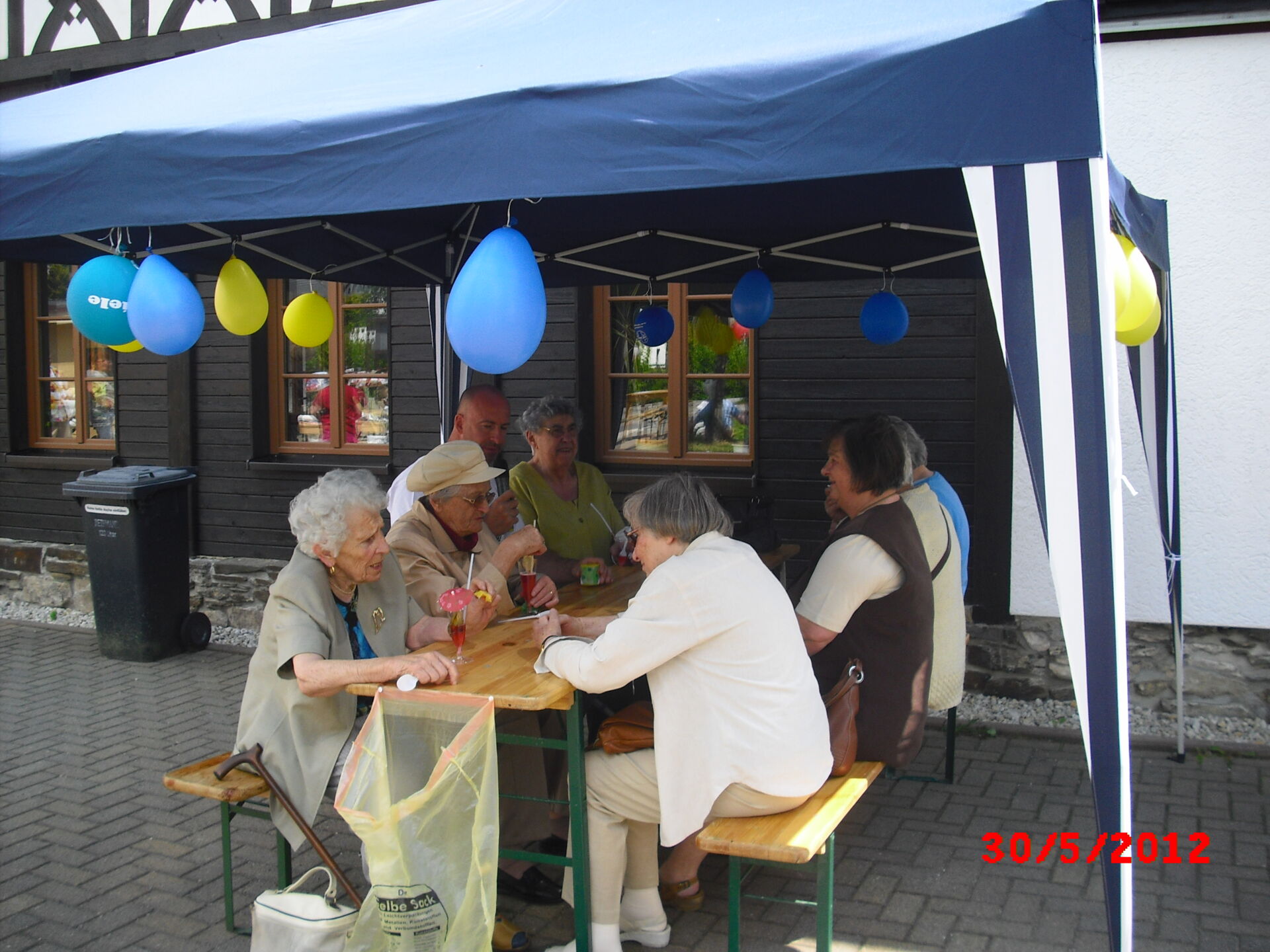 Eine Gruppe älterer Menschen sitzt fröhlich unter einem Pavillon mit bunten Ballons. Feier der Hauskrankenpflege Sylke Bauer in Zschorlau.
